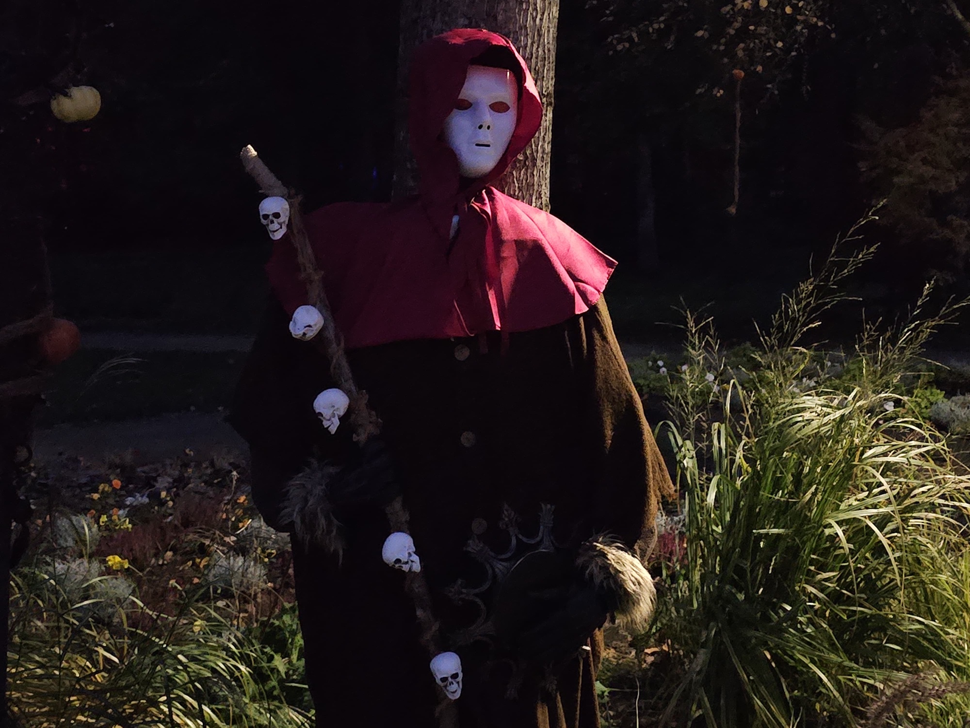 Gestalt mit roter Kapuze, Maske und Stab mit Totenköpfen